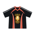 A black short-sleeve V-neck athletic-style shirt with coral orange shoulder and side panels, white stripes on the sleeves, and a large golden-yellow graphic print on the chest reading 'THE DOOR WAS NEVER LOCKED' with additional text below.